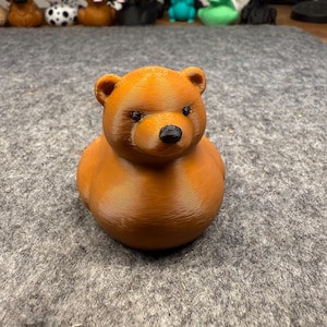May include: A brown bear-shaped rubber duck with a black nose and eyes. The duck is the focal point, with other animal-shaped ducks in the background. The duck is made of a 3D printed material.