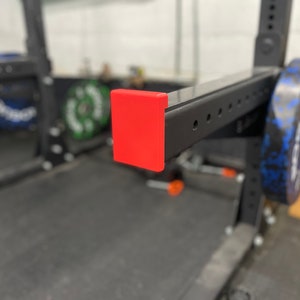 May include: Close-up of a red plastic end cap on a black metal weightlifting rack. The rack has a silver metal bar and a blue and black weight plate.
