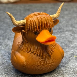 Puede incluir: Un pato de goma marrón con un diseño de vaca de las Tierras Altas. El pato tiene un pico naranja y cuernos color crema. La cabeza del pato presenta pelo detallado y pequeños ojos negros. Es un artículo de novedad.