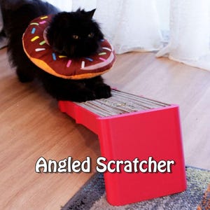 May include: A black cat wearing a donut-shaped recovery collar stands on a red angled cat scratcher. The scratcher has a cardboard scratching surface. The text "Angled Scratcher" is displayed on the red base.