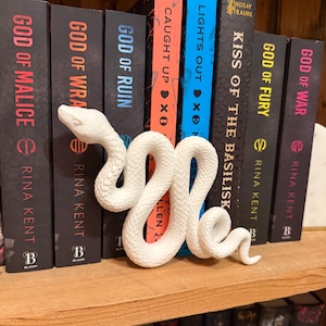 May include: A white snake bookend coiled in front of a row of books on a wooden shelf. The book titles include "God of Malice," "God of Wrath," and "Kiss of the Basilisk." The books have black covers with various accent colors.