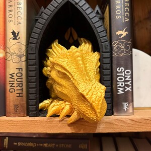 May include: A golden dragon head emerges from a black gothic archway, placed on a wooden shelf between two books. The books are titled "Fourth Wing" and "Onyx Storm" by Rebecca Yarros.
