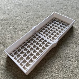 May include: A white rectangular tray with a patterned grid design. The tray has a series of interconnected, curved shapes forming the grid. The tray is made of a solid, opaque white material and is designed to hold small items.