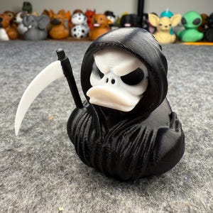 Puede incluir: Una figura de pato impresa en 3D en blanco y negro vestida como la Parca, completa con una capa con capucha y una guadaña. El pato tiene una cara de calavera y forma parte de una colección de figuras novedosas.