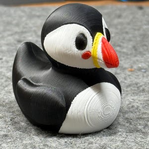 May include: A 3D printed puffin figurine featuring a black back, wings, and head, with a white chest and face patches. The bird has a bright red beak with a yellow base, black eyes, and small red circular markings on its cheeks, showing visible layer lines.