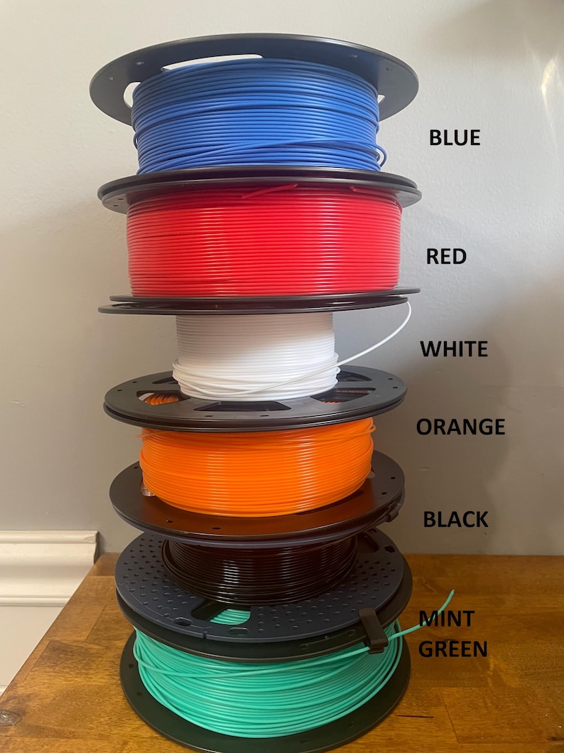 May include: A stack of six spools of filament for 3D printing. The spools are arranged from top to bottom in the following colors: blue, red, white, orange, black, and mint green.