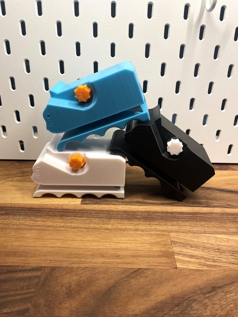 May include: Three 3D printed clamps, one blue, one white, and one black, with orange and white knobs. The clamps are stacked on top of each other on a wooden surface.