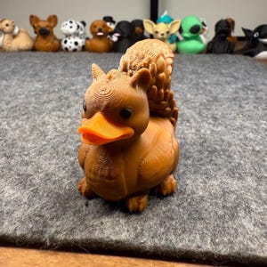 May include: A brown duck-squirrel hybrid figurine with an orange beak and detailed texture. Other animal figurines are visible in the background. The figurine is on a gray surface.