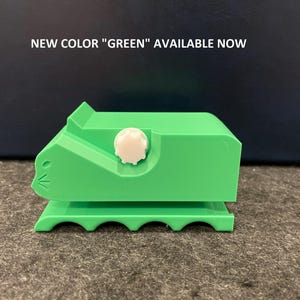 May include: A green plastic tape dispenser shaped like a cat with a white knob. The dispenser has a scalloped base and the text "NEW COLOR "GREEN" AVAILABLE NOW" is visible in the background.