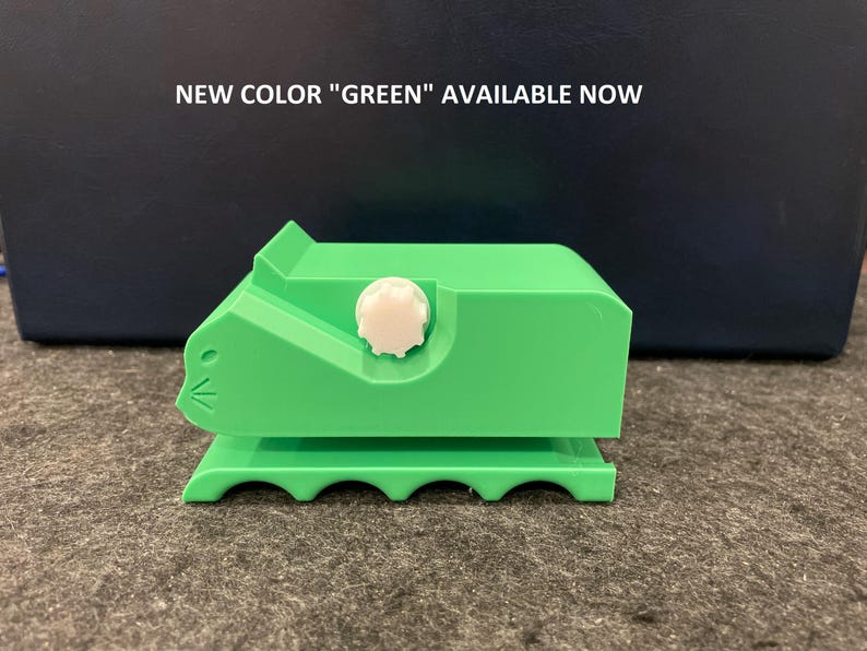 May include: A green plastic tape dispenser shaped like a cat with a white knob. The dispenser has a scalloped base and the text "NEW COLOR "GREEN" AVAILABLE NOW" is visible in the background.