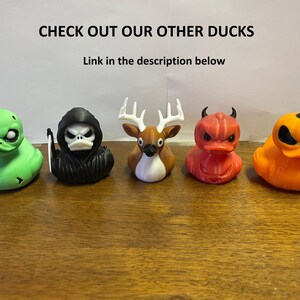 May include: Five rubber duck toys with different Halloween themes. The ducks are green, black, brown, red, and orange. The ducks are all facing forward and have different expressions on their faces. The text "CHECK OUT OUR OTHER DUCKS" is at the top of the image. The text "Link in the description below" is below the first line of text.