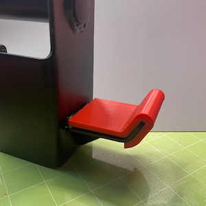 Könnte beinhalten: Eine schwarze Wandhalterung aus Metall mit einem roten Haken aus Kunststoff. Der Haken ist für das Aufhängen einer Langhantel konzipiert.