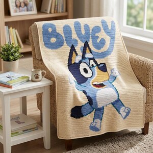 Pode incluir: Um cobertor de croch&ecirc; bege com a palavra "Bluey" em letras azul claro e uma imagem do personagem de desenho animado Bluey. O cobertor est&aacute; sobre uma poltrona de vime. Uma pequena mesa lateral branca tem uma caneca e livros.