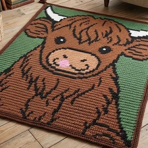 May include: A handmade crochet rug featuring a Highland cow design. The rug showcases a brown cow with black accents, white horns, and a pink tongue, set against a green background. The border is a darker brown. This decorative rug adds a touch of whimsy to any room.