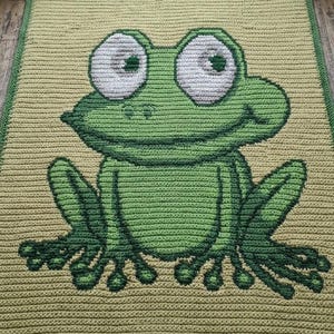 May include: A rectangular crochet blanket featuring a large, centered green frog graphic. The frog has prominent white eyes with dark green pupils, a wide smile, and splayed feet. The background is a light olive green, framed by a dark green border.