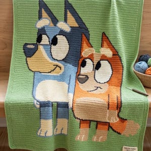 Op de afbeelding: Een gehaakte deken met twee cartoonhonden in blauw, oranje en bruin op een groene achtergrond. De deken heeft een getextureerd uiterlijk en een rand. Het label luidt "HEELER FAMILY CROCHET".