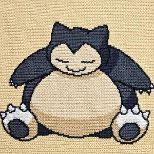 May include: A crochet blanket featuring a large, centered graphic of the character Snorlax. The character is depicted in dark blue, cream, and white yarn, set against a solid light beige background. The stitches are uniform and tightly woven.