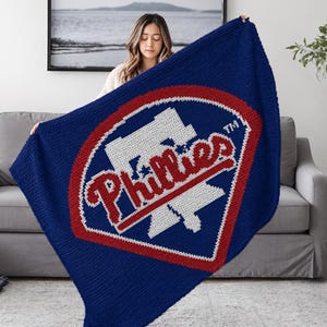 May include: A blue blanket featuring the Philadelphia Phillies logo in red and white. The logo is a stylized baseball with the team name in a cursive font. The blanket is made of a soft, knitted material.