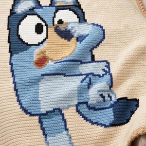 Könnte beinhalten: Eine gehäkelte Decke mit einer Cartoon-Hundefigur in Blau- und Hellblautönen auf beigem Hintergrund. Die Decke hat ein detailliertes Design mit einem beigen Rand und einem strukturierten Aussehen, geeignet für ein Kinderzimmer oder einen Kindergarten.