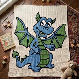 May include: A cream-colored crocheted blanket featuring a pixelated blue and green dragon design. The dragon has large wings and a friendly expression. A children's book and a teddy bear are nearby.