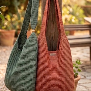 Pode incluir: Duas bolsas de ombro de crochê feitas à mão. Uma é verde suave, a outra é de cor coral quente. Ambas as bolsas têm alças longas e formato arredondado. As bolsas estão penduradas em um poste de madeira, sugerindo um estilo rústico ou boêmio.