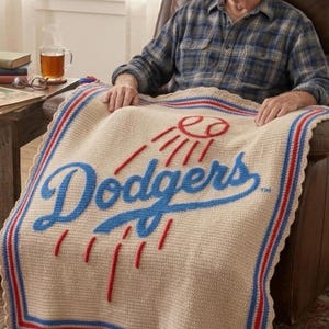 May include: A beige crocheted blanket featuring the word "Dodgers" in blue script, with a red and blue border. The blanket also has a red baseball and sunburst design. The blanket is draped over a chair.