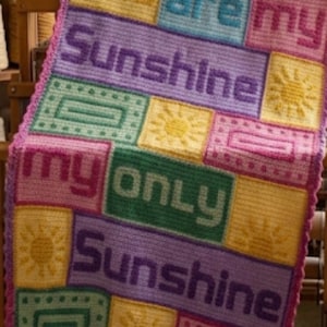 Op de afbeelding: Een veelkleurige gehaakte deken met de tekst "you are my Sunshine my only Sunshine" in blokletters. Het ontwerp bevat afwisselende rechthoekige panelen in geel, blauw, roze, paars en groen, voorzien van zonmotieven en geometrische randen.