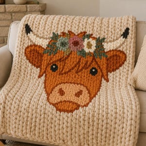 May include: A chunky knit cream-colored throw blanket featuring a large, centered graphic of a highland cow face. The cow is rendered in shades of rust orange and brown, adorned with a floral crown of muted pink, blue, and white yarn flowers.