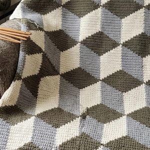 May include: A crocheted blanket featuring a repeating geometric pattern of cubes in shades of olive green, light gray, and cream. The textile is constructed with a dense, textured stitch pattern that creates a three-dimensional visual effect.