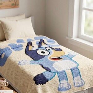 Pode incluir: Um cobertor de croch&ecirc; bege e azul com um personagem de desenho animado de cachorro com corpo azul, manchas pretas e focinho amarelo. O cobertor est&aacute; em uma cama com travesseiros brancos.