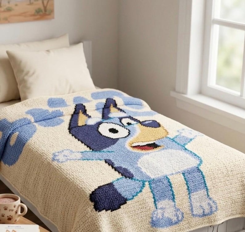 Pode incluir: Um cobertor de croch&ecirc; bege e azul com um personagem de desenho animado de cachorro com corpo azul, manchas pretas e focinho amarelo. O cobertor est&aacute; em uma cama com travesseiros brancos.