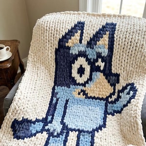 Puede incluir: Una manta de punto grueso con un gráfico grande de estilo pixelado de un perro azul. La manta es principalmente de color crema con hilos azul oscuro, azul claro y amarillo pálido utilizados para crear la figura central del perro.
