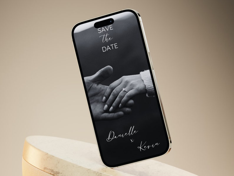 Puede incluir: Una foto en blanco y negro de las manos de una pareja con un anillo en el dedo de la mujer. La foto se muestra en la pantalla de un tel&eacute;fono inteligente con el texto "Save the Date" y "Danielle & Kevin" en la pantalla.
