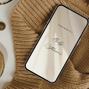 May include: A smartphone displaying a digital invitation to a baby shower. The invitation reads "You're invited to a Baby Shower". The phone is lying on a brown knitted sweater.