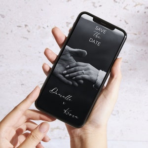 Puede incluir: Una foto en blanco y negro en la pantalla de un tel&eacute;fono inteligente muestra dos manos entrelazadas con un anillo en un dedo. El texto "Save the Date" est&aacute; en la parte superior de la pantalla y el texto "Danielle & Kevin" est&aacute; en la parte inferior de la pantalla.