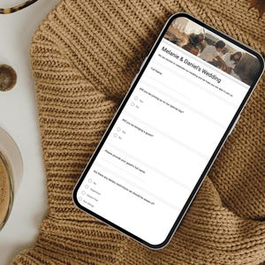 May include: A smartphone showing a wedding invitation form, with the text "Melanie & Daniel's Wedding". The phone is placed on a tan knitted jumper, with a cup of coffee and eyeglasses in the background.