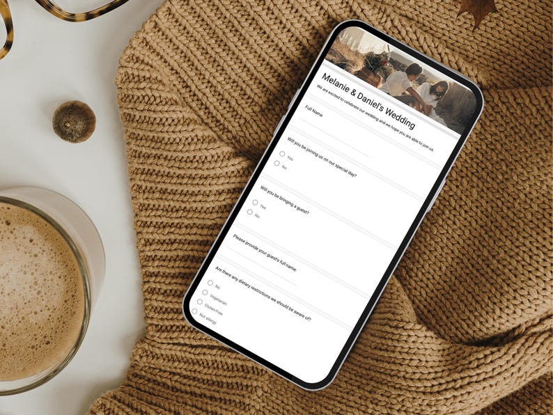 May include: A smartphone showing a wedding invitation form, with the text "Melanie & Daniel's Wedding". The phone is placed on a tan knitted jumper, with a cup of coffee and eyeglasses in the background.