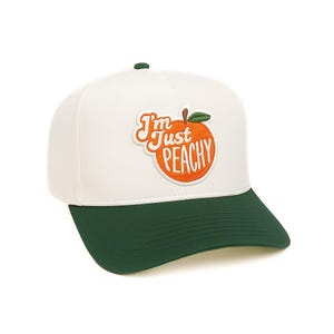 May include: White and green baseball cap with a peach graphic. The cap features the phrase "I'm Just Peachy" in orange and white lettering, with a peach illustration. The brim and the top button are green.