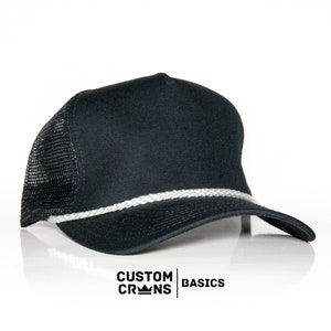 May include: Black trucker hat with a white rope trim. The hat has a black mesh back and a black front panel. The text "CUSTOM CROWNS BASICS" is printed on the front of the hat.