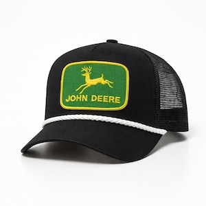 May include: Black trucker hat with a green and yellow John Deere logo patch. The patch features a leaping deer. The hat has a white rope detail and a mesh back.