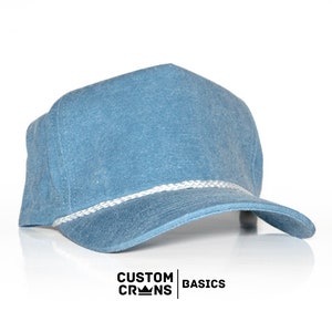 May include: A blue denim baseball cap with a white rope trim. The cap has a slightly distressed look and features the text "CUSTOM CROWNS BASICS" on the front.