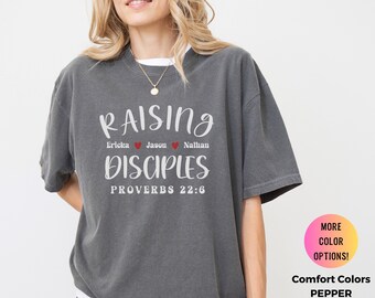 May include: A dark gray t-shirt with the words "RAISING DISCIPLES" in white script. The names Ericka, Jason, and Nathan are listed with hearts. The text "PROVERBS 22:6" is below. The shirt is Comfort Colors, Pepper.
