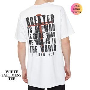 Könnte beinhalten: Weißes, langes T-Shirt mit dem Text "GREATER IS HE WHO IS IN ME THAN HE WHO IS IN THE WORLD 1 JOHN 4:4" auf dem Rücken. Der Text ist schwarz mit einem roten Kreuz-Design.
