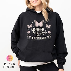 May include: Black hoodie with the text "MOTHERHOOD IS MY MINISTRY" and a butterfly and floral design. The hoodie also has the text "PROVERBS 22:6". The image also has the text "MORE COLOR OPTIONS!" and "BLACK HOODIE".