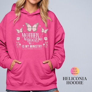 May include: A pink hoodie with the text "MOTHERHOOD IS MY MINISTRY" and butterfly graphics. The hoodie has a drawstring hood and a front pocket. The text "PROVERBS 22:6" is below the main text. The words "MORE COLOR OPTIONS!" and "HELICONIA HOODIE" are also visible.