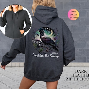 May include: Dark heather zip-up hoodie with a graphic of a raven perched on a branch, set against a night sky with a crescent moon and stars. The text "Consider The Ravens" is below the image. The hoodie has a front pocket and a hood.