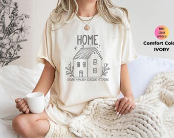 May include: A woman wearing a cream colored t-shirt with a black and white illustration of a house and the text "HOME GROWING MAKING SCHOOLING STEADING". The shirt is on a white background.