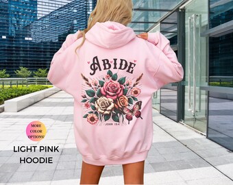 May include: Light pink hoodie with a floral design and the word "ABIDE" in black lettering. The design also includes the text "JOHN 15:4".