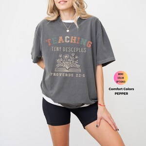 May include: A gray t-shirt with a vintage-style graphic that says "Teaching Tiny Disciples Proverbs 22:6". The graphic features a book with flowers growing out of it. The shirt is available in other colors.