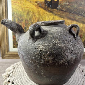 Könnte beinhalten: Eine rustikale braune Tonkanne mit zwei Henkeln und einem Ausguss. Die Kanne hat eine raue, strukturierte Oberfläche und scheint sehr alt zu sein.